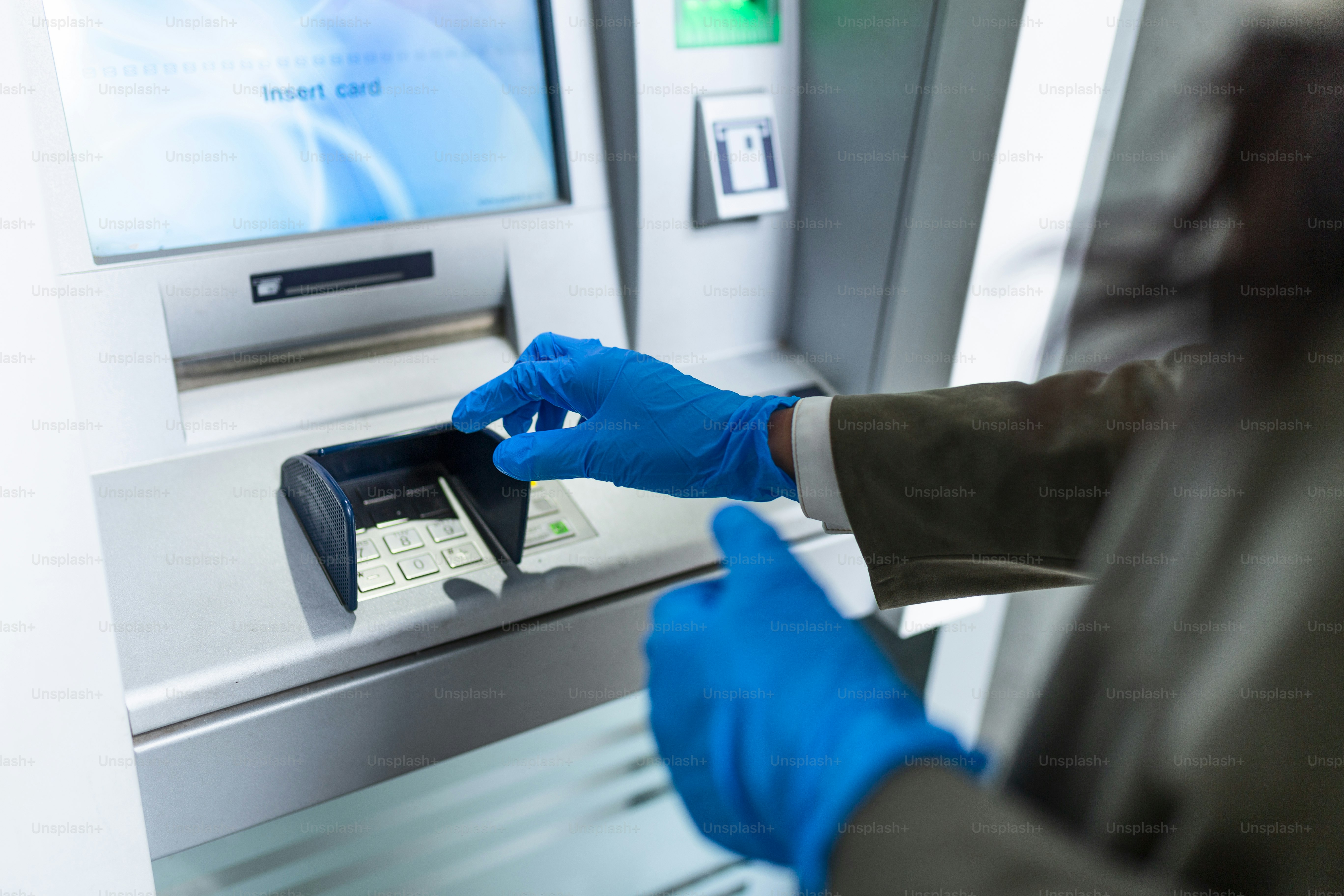 Giovane donna con maschera protettiva e guanti in piedi sulla strada della città e utilizzando il bancomat per prelevare contanti. Concetto di pandemia di corona o virus Covid-19.