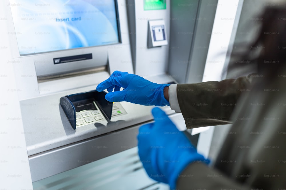 Young woman with protective mask and gloves standing on city street and using ATM machine to withdraw cash. Corona or Covid-19 virus pandemic concept.