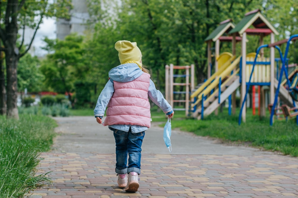 Little girl walking to the kids playground holding protective face mask after the end of quarantine. Pandemic COVID-19 is over concept.