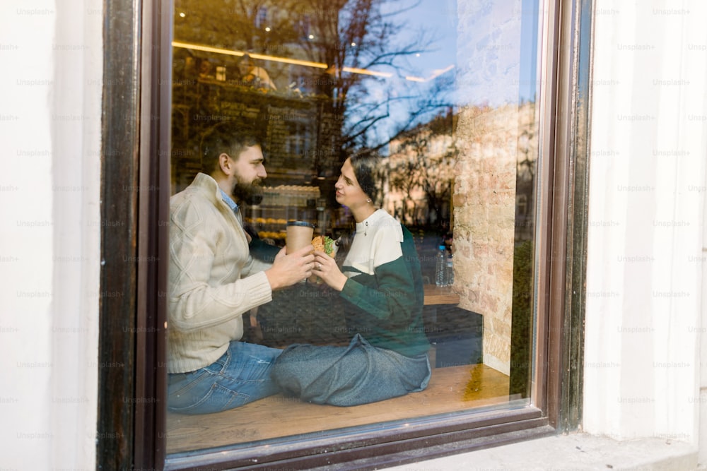 Joven pareja romántica, mirándose, bebiendo café y comiendo croissants franceses tradicionales en una acogedora cafetería. Disparado a través de la ventana desde la calle.
