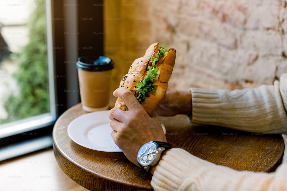 Close up cropped image of male hands holding croissant stuffed with lettuce and fresh vegetables. Cafe concept. Healthy food. Lunch and breakfast concept. Coffee to go on the background.
