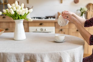 Cropped view of woman pouring beverage in cup. Kitchen with wooden interior style, white spring flowers and tablecloth on dinner table
