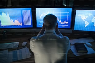 Rückansicht eines frustrierten Geschäftsmannes mit Computer, der am Schreibtisch sitzt und spät arbeitet. Finanzkrisenkonzept.