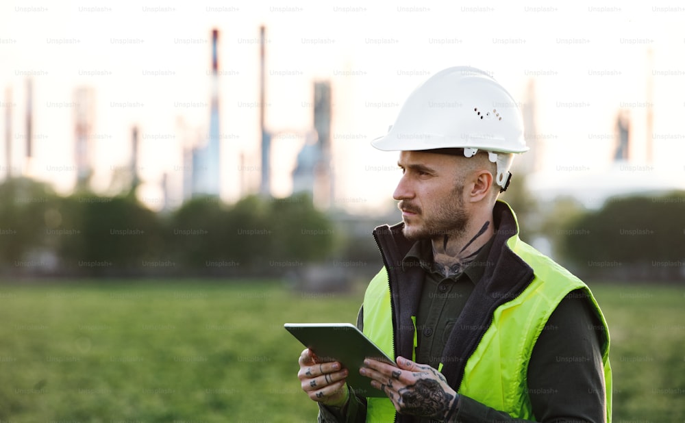 Serious young engineer with tablet standing outdoors by oil refinery. Copy space.
