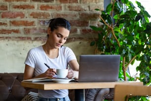 Beautiful young girl working at a coffee shop with a laptop and taking notes. Female freelancer connecting to internet via computer. Business woman indoor with coffee and laptop taking notes