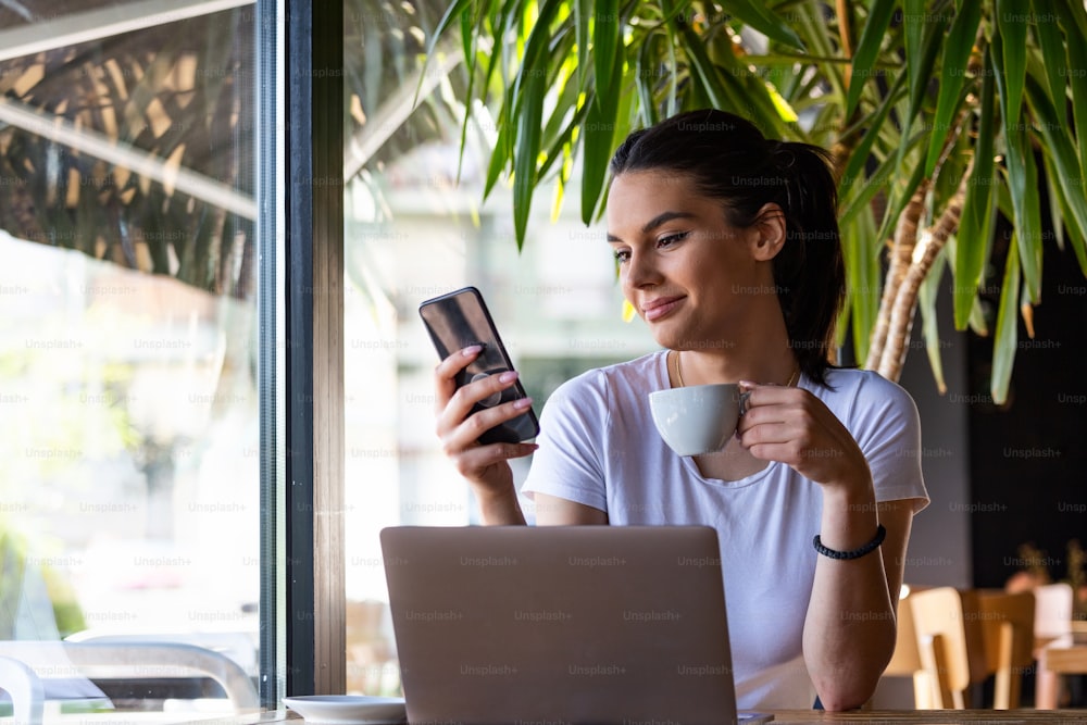 Smiling woman drinking coffee and using her mobile phone. Satisfied female enjoying cup of coffee wile working from coffee shop. Close up portrait of beautiful girl drinking coffee in the coffee shop