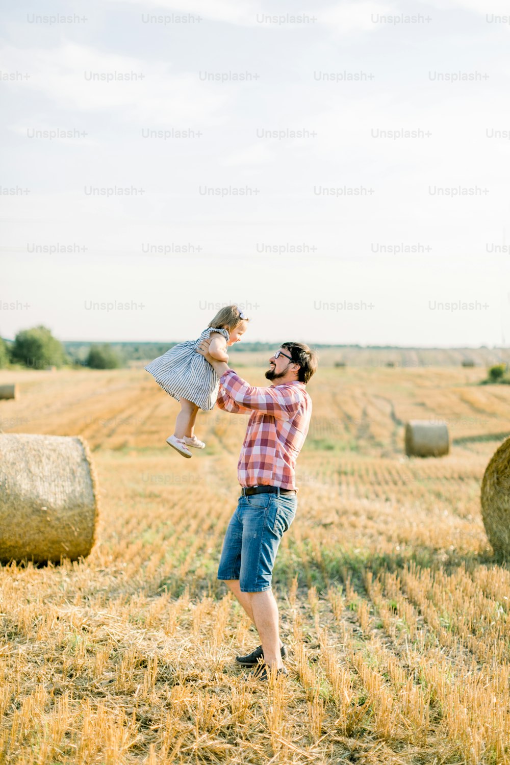Handsome young bearded father dancing with his little cute daughter, holding her on hands, having fun in summer sunset field with hay bales on the background. Outdoor summer family shot.