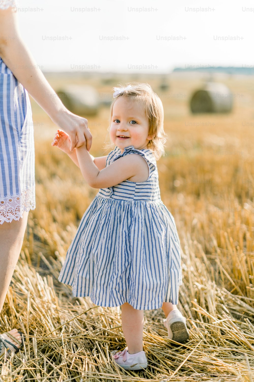 Emotional portrait of a happy and joyful little girl in striped dress, holding hand of her mom while walking together in a wheat field with hay bales in the rays of the sunset. Summer. Childhood.