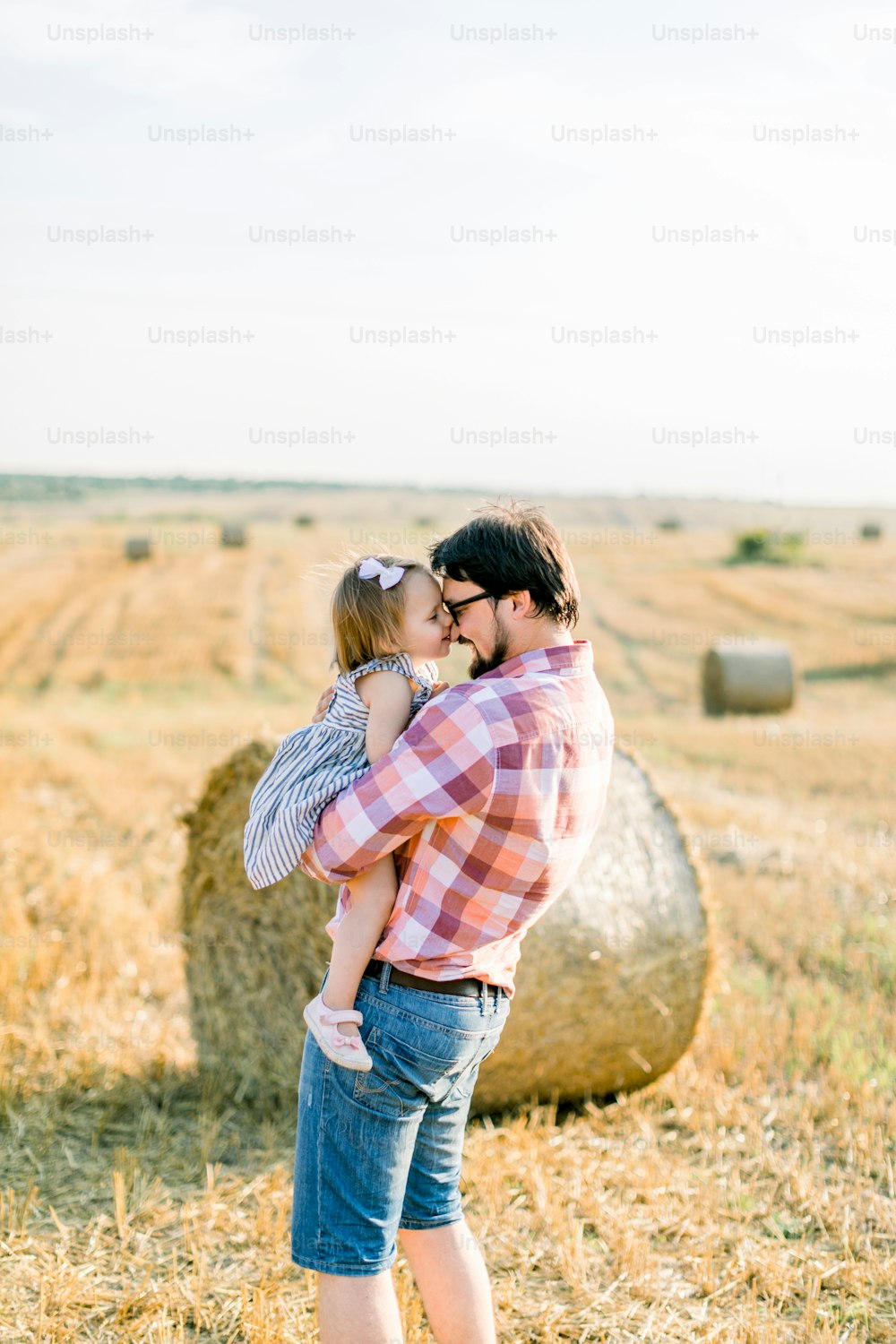 Happy joyful young father, handsome bearded man in jeans and checkered shirt, holding on hands his pretty little baby girl in dress, hugging and touching foreheads, walking in field next to hay bales.