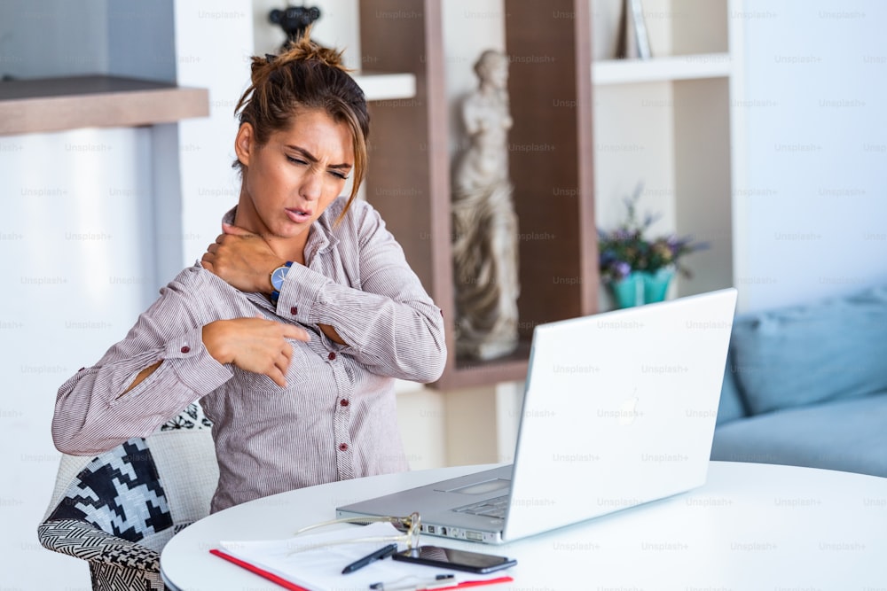 Retrato da jovem mulher estressada sentada na mesa do escritório em casa na frente do laptop, tocando as costas doloridas com expressão dolorida, sofrendo de dor nas costas depois de trabalhar no pc