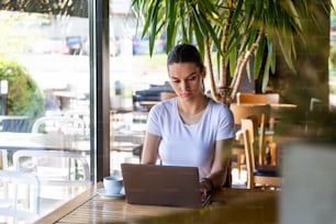 Young beautiful girl sitting at a table in a cafe by the window, drinking cappuccino coffee. Beautiful young working with laptop from coffee shop. Attractive woman sitting in a cafe with a laptop