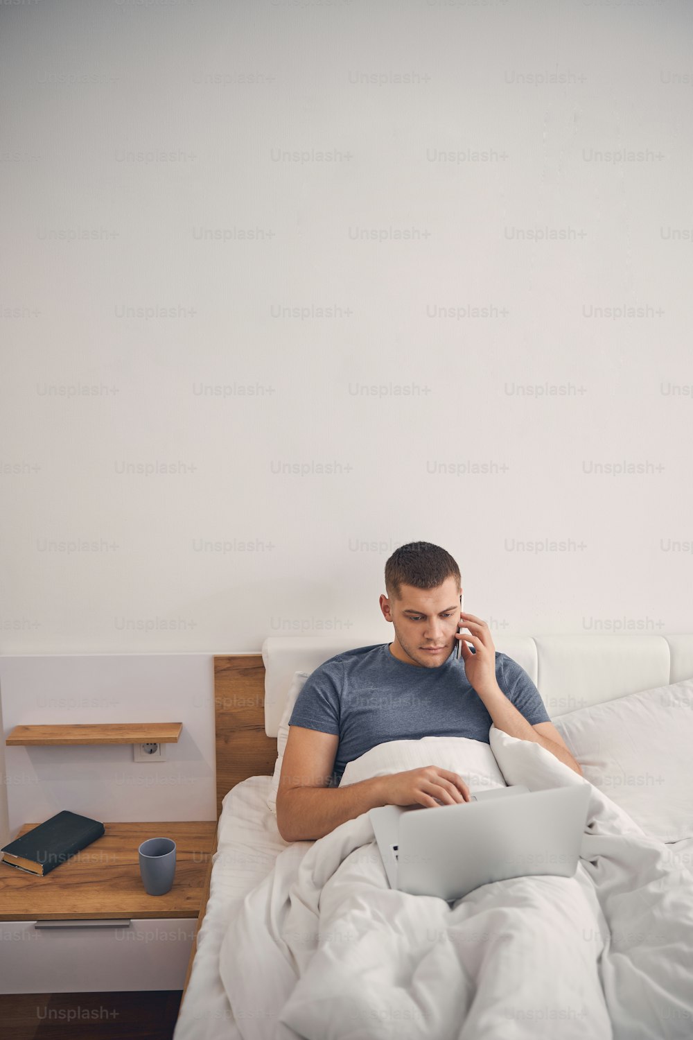 Attractive brunette male talking on phone while staying in bed with computer