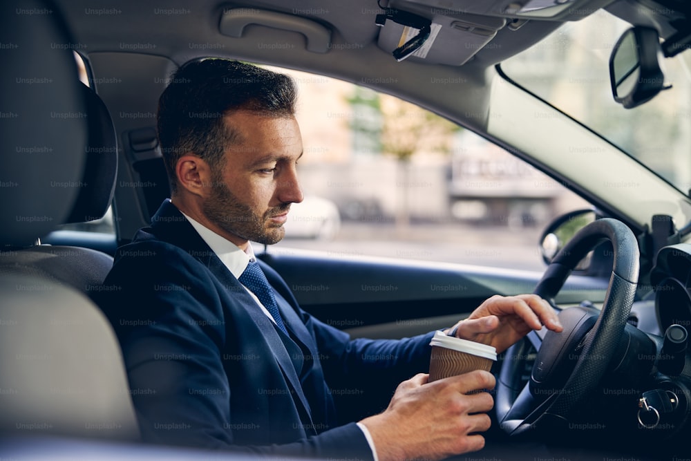 Attractive brunette businessman in official clothes holding coffee while driving auto