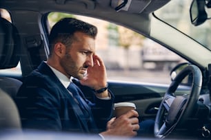 Handsome brunette man in formal clothes with coffee in hand touching head while driving