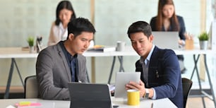 Businessmen are working together with computer tablets while sitting at the white working desk over employees as background.
