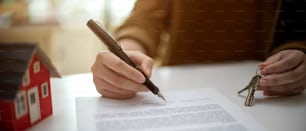 Close up view of a woman signing home loan agreement while holding house key on white table with house model