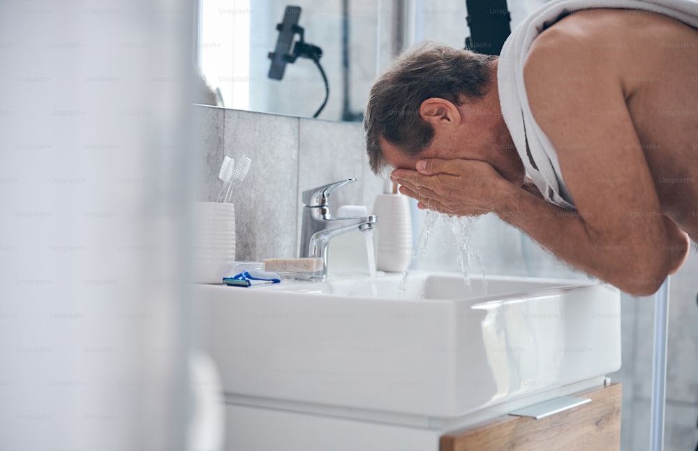 Side view of a male with a towel over his shoulder leaning over the sink