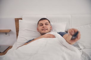Handsome Caucasian male in blue T-shirt smiling and relaxing in bed while stretching his hand