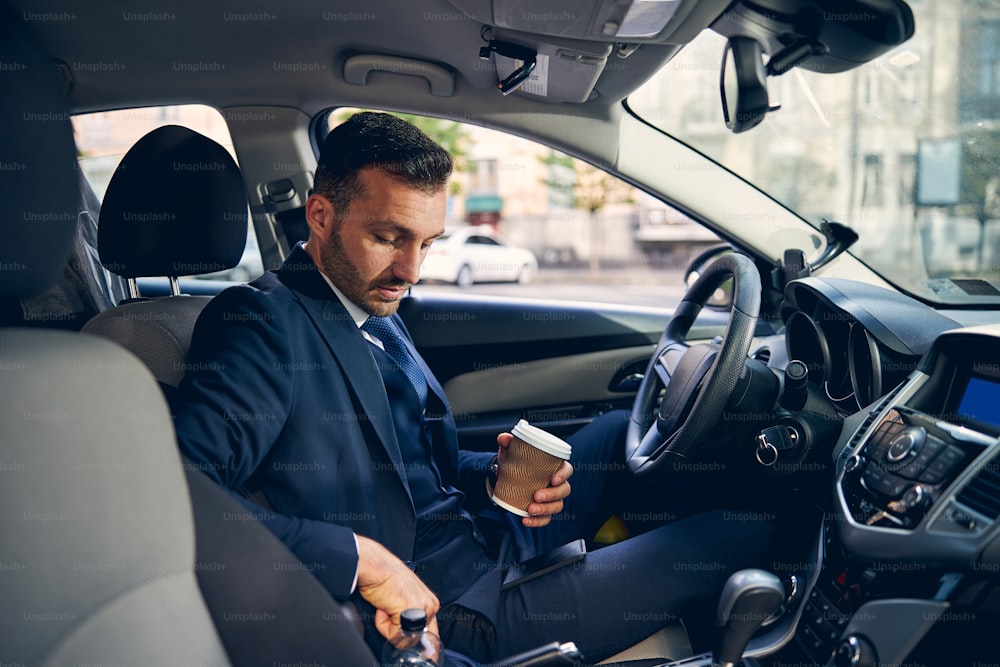 Successful brunette businessman in trendy suit spending time in car while holding cup of coffee