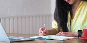 The young woman is taking notes while sitting in front of her computer laptop at the wooden working desk.