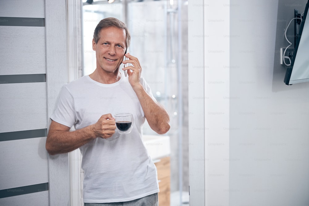 Front view of a pleased male with a cup of coffee calling on the phone