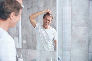 Waist-up portrait of a joyous middle-aged handsome Caucasian man styling his hair in the bathroom