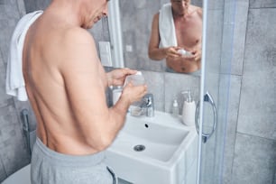Fokussierter Mann ohne Shirt mit einem Badetuch über der Schulter, der vor dem Keramikwaschbecken steht