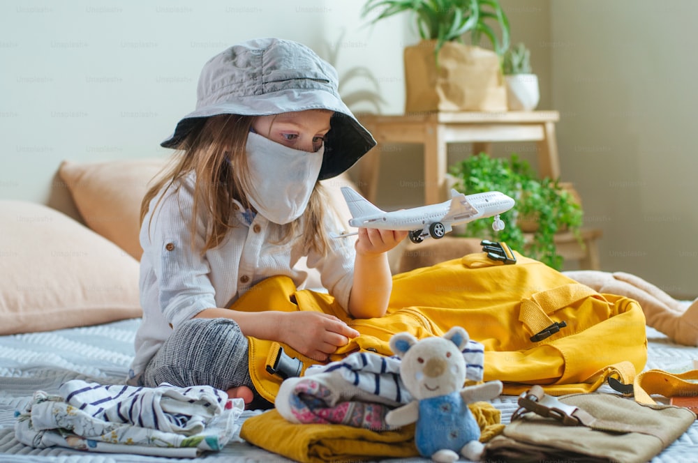 Little toddler girl in protective face mask playing with airplane toy packing her backpack for the travel sitting on the sofa indoor. Prepairing for a travel after the end of quarantine. New life after Pandemic COVID-19 concept.