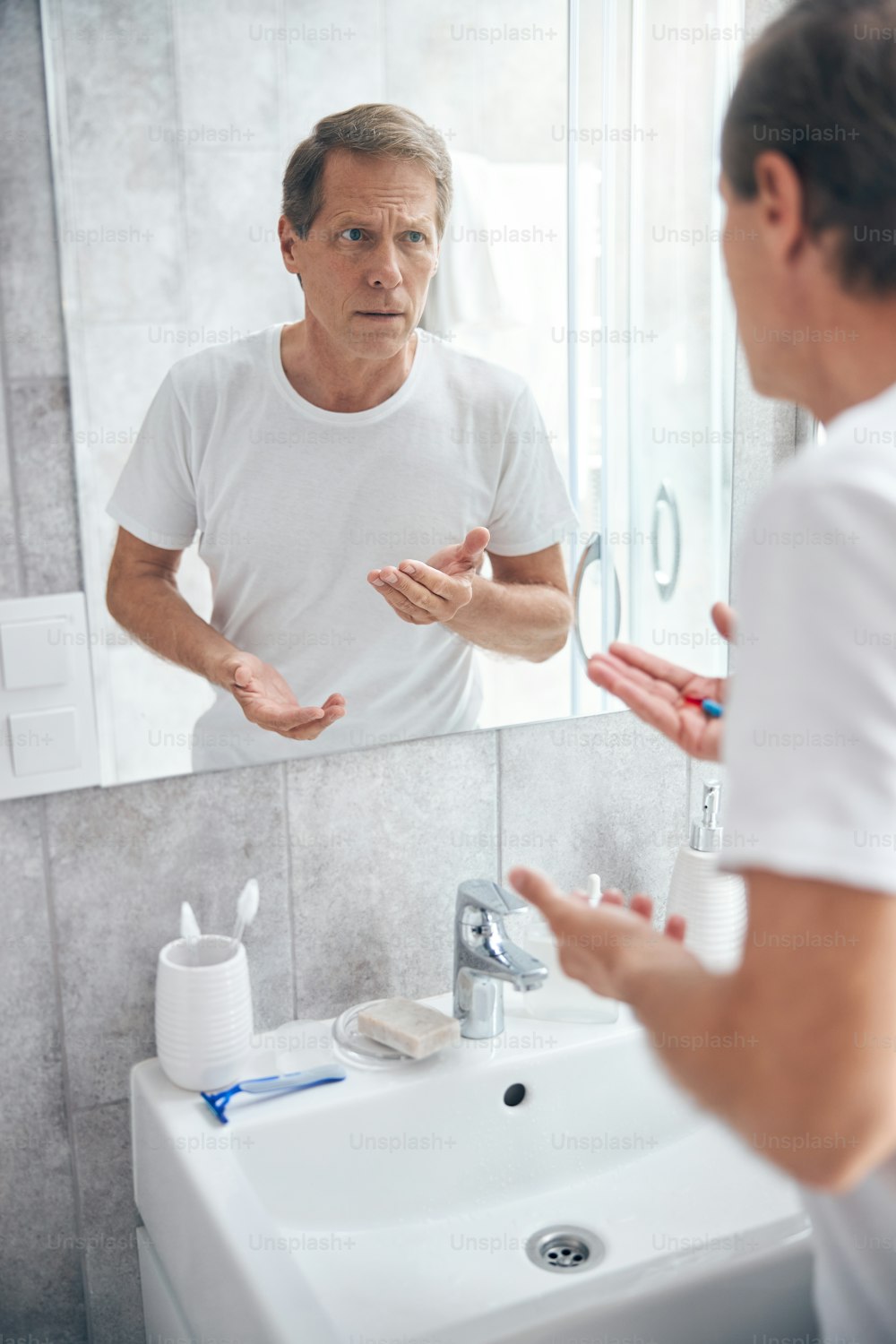 Serious male with a pair of capsules in his hand staring in the bathroom mirror
