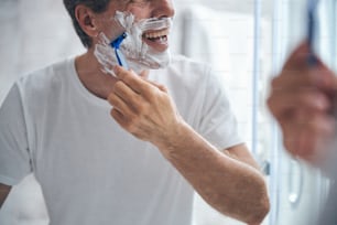 Foto recortada de un alegre hombre caucásico de pelo corto afeitándose la cara con una maquinilla de afeitar