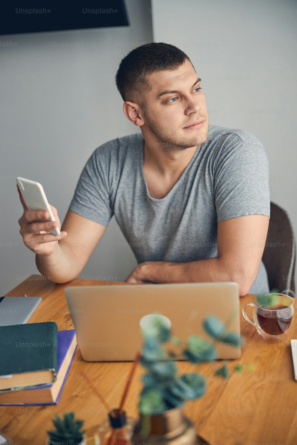 Concentrated brunette male with blue eyes working online from home while holding mobile in hands and contemplating