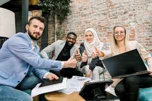 Successful excited multi-ethnic group of business people sitting together around the table with papers and laptop, showing thumbs up gesture, looking at camera and smiling.
