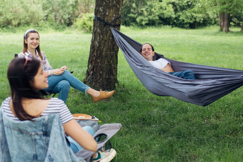 Social distancing. Small group of people enjoying conversation at picnic with social distance in summer park. Friends chilling in hammock and on chairs among trees. Safety gatherings in new world
