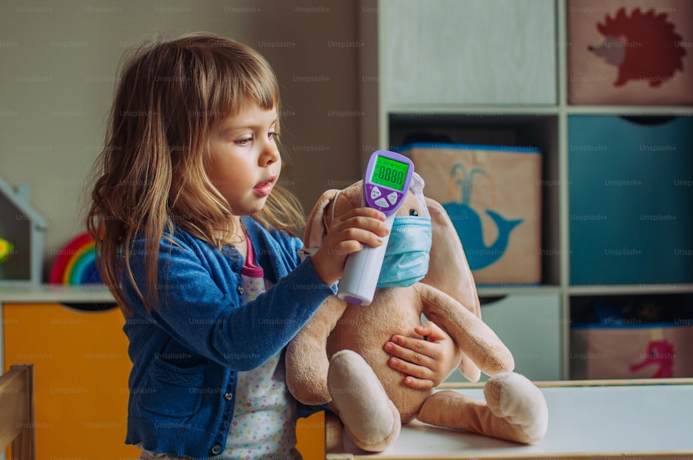 Kleinkind mit elektronischem berührungslosem Thermometer für Hasentier im Kinderzimmer. Pandemie-COVID-19-Konzept.