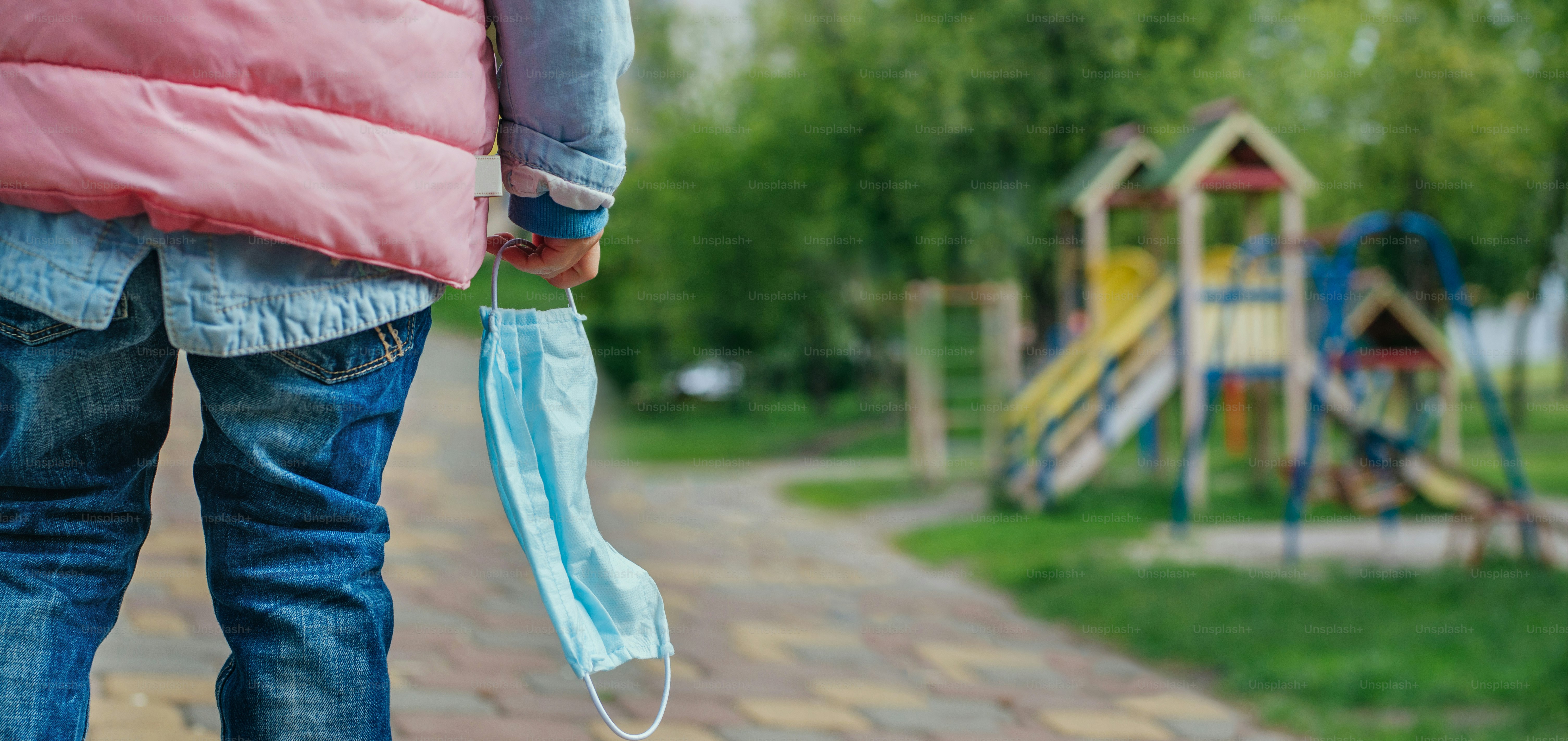 Close up of little girl take off and holding protective face mask looking at the open playground after the end of quarantine. Pandemic COVID-19 is over concept. Pnoramic background.