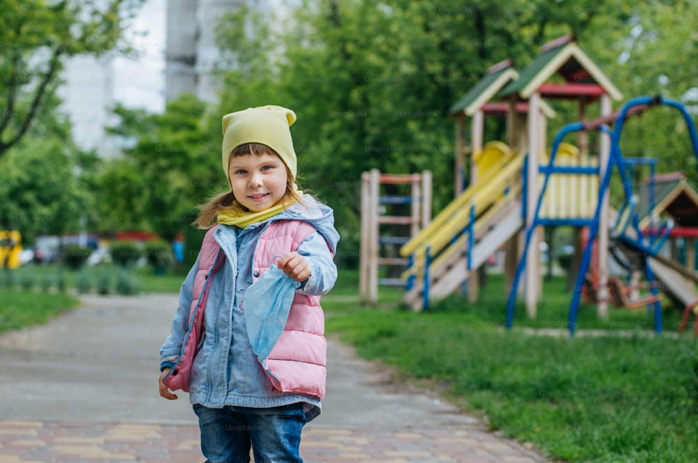 Happy little girl holding protective face mask near open playground after the end of quarantine. Pandemic COVID-19 is over concept.