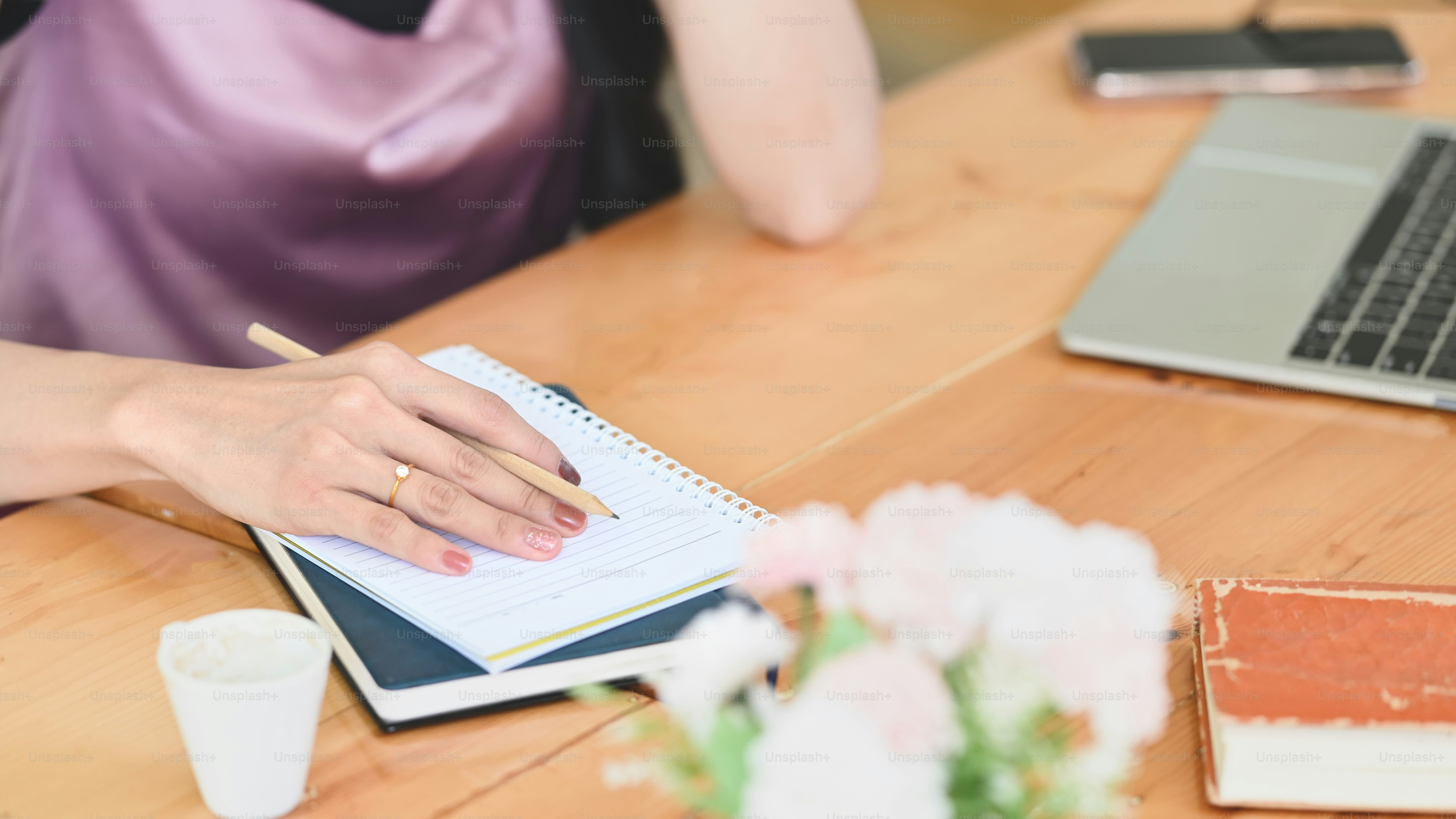 A woman is writing on a notebook while sitting at the wooden working desk.