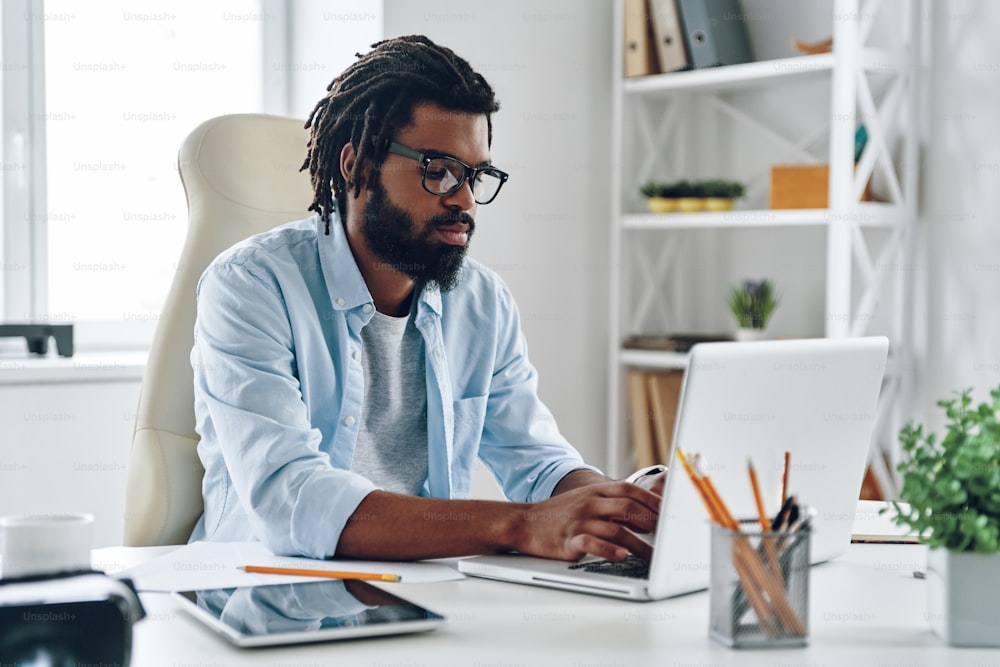 Nachdenklicher junger afrikanischer Mann in Brillen, der während der Arbeit im Büro am Computer arbeitet