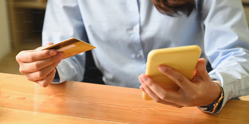 Imagen recortada de una mujer sostiene una tarjeta de crédito y un teléfono inteligente en las manos mientras está sentada en la sala de estar.