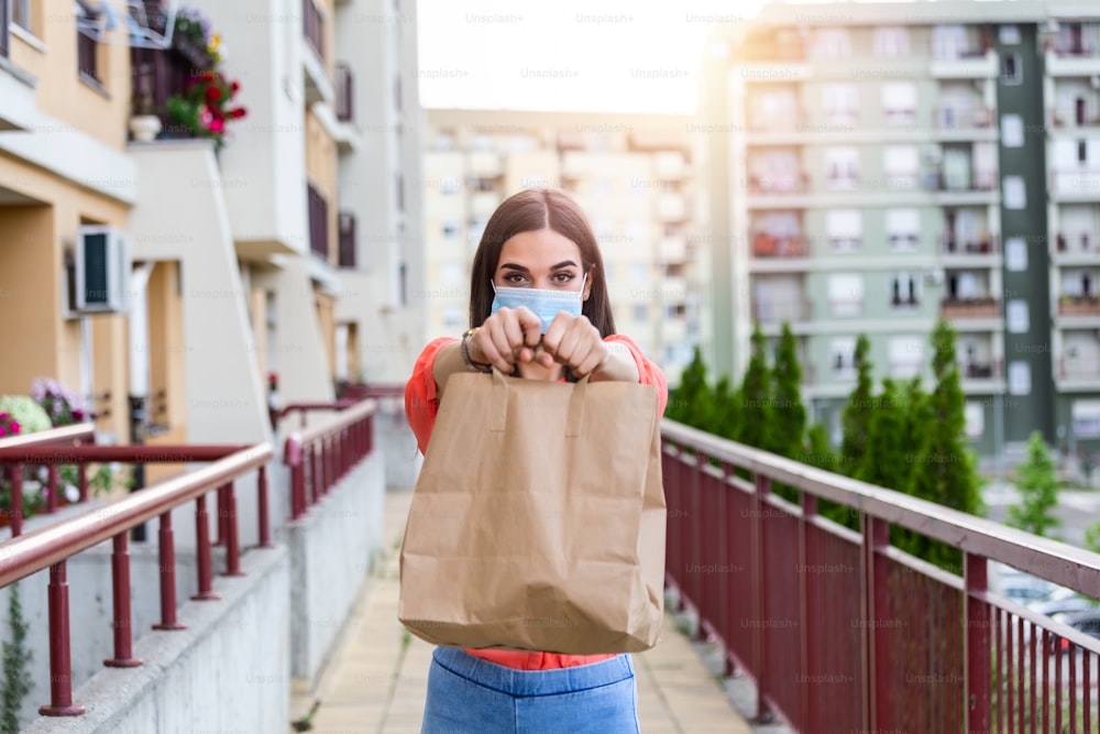 Contactless delivery of goods during Covid 19, coronavirus pandemic. The food delivery courier is holding a large paper bag in her hands. Woman with medical mask delivering food.