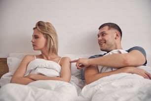 Cute blond woman looking angry in bed while brunette male trying to make her smile