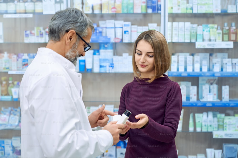 Pharmacist offering pretty female customer medical products in drugstore. Young woman holding white bottles, choosing product. Side view of mature chemist wearing glasses and white lab coat.