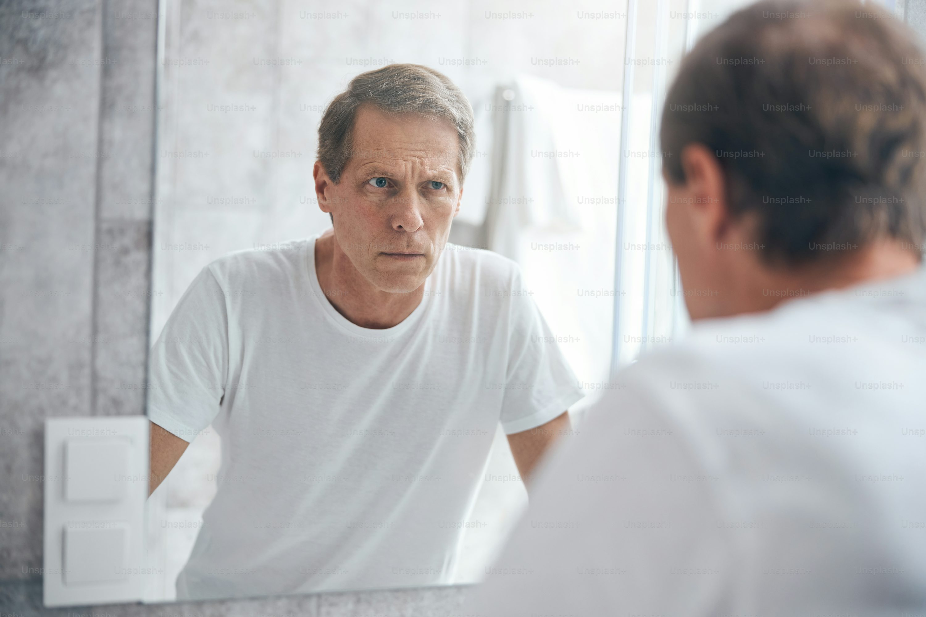 Waist-up portrait of a sulky Caucasian man in a t-shirt staring fixedly at his appearance