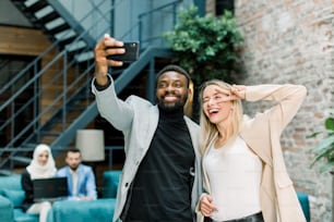 Gente de negocios feliz, trabajando en la oficina. Mujer caucásica sonriente y hombre africano haciendo una foto selfie conjunta en el teléfono inteligente, posando, riendo y mostrando gestos dofferent.