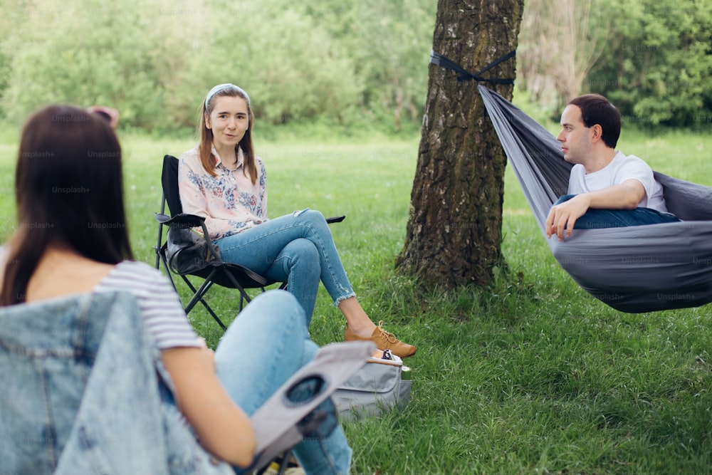 Social distancing. Small group of people enjoying conversation at picnic with social distance in summer park. Friends chilling in hammock and chairs among trees. New normal, safety gatherings