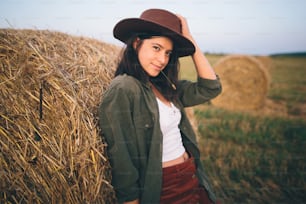 Countryside slow life. Stylish happy girl posing at hay bale in summer field in sunset. Portrait of young sensual woman in hat smiling at haystack, atmospheric tranquil moment.
