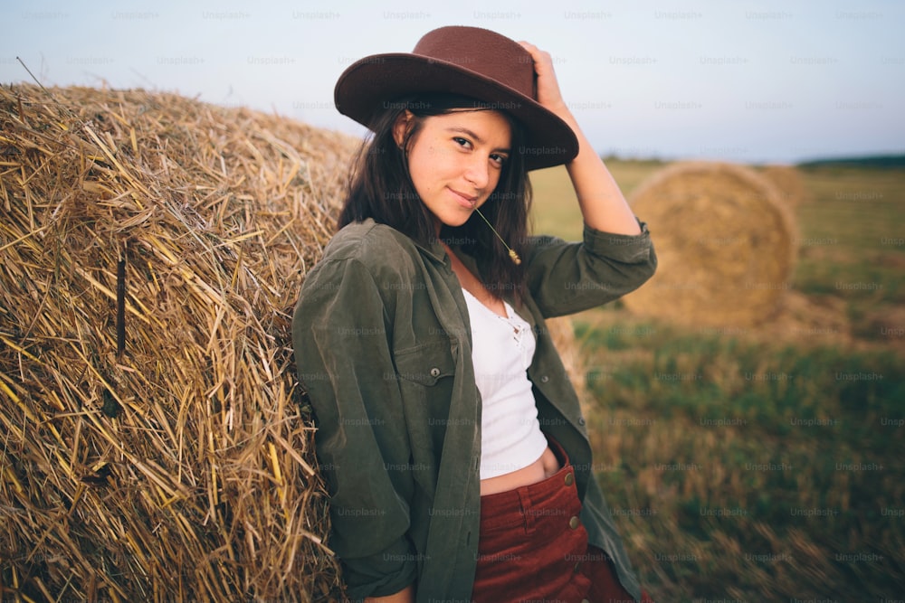 Countryside slow life. Stylish happy girl posing at hay bale in summer field in sunset. Portrait of young sensual woman in hat smiling at haystack, atmospheric tranquil moment.
