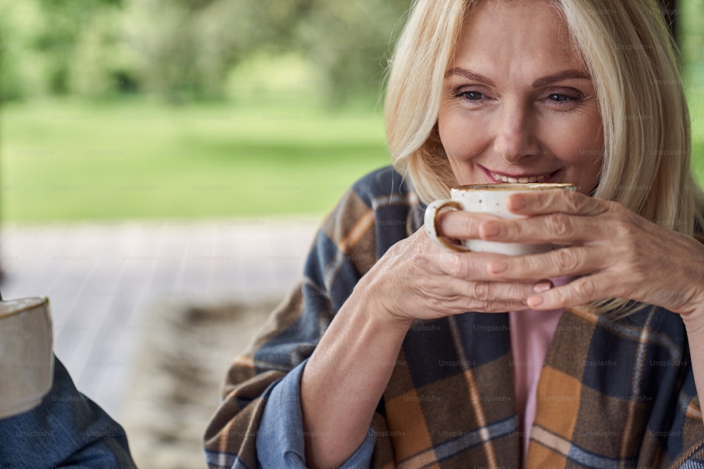 웃고 있는 매력적인 성숙한 여성은 녹색 자연 가운데 집의 열린 테라스에서 차를 마시고 있다