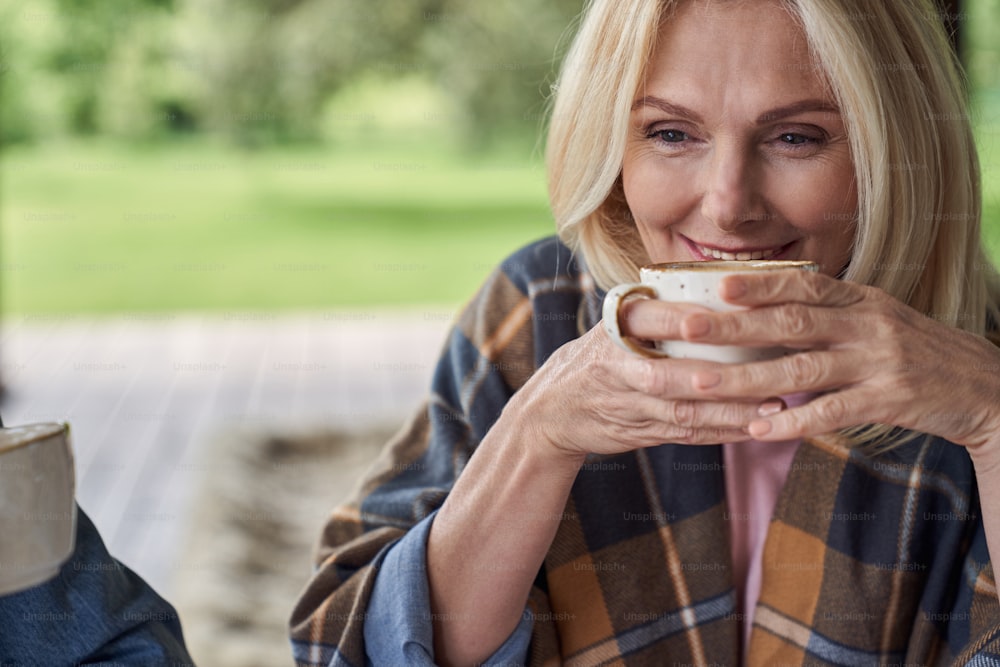 Smiling attractive mature female is drinking tea in open terrace of house among green nature