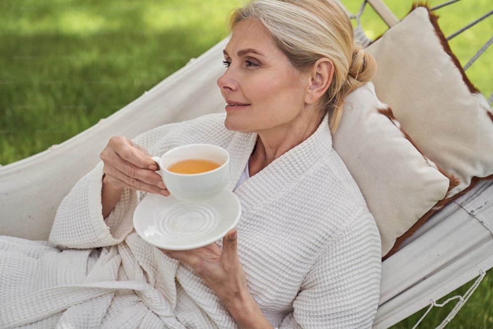 陽気な成熟した女性がハンモックに横たわり、屋外で温かい飲み物を飲む